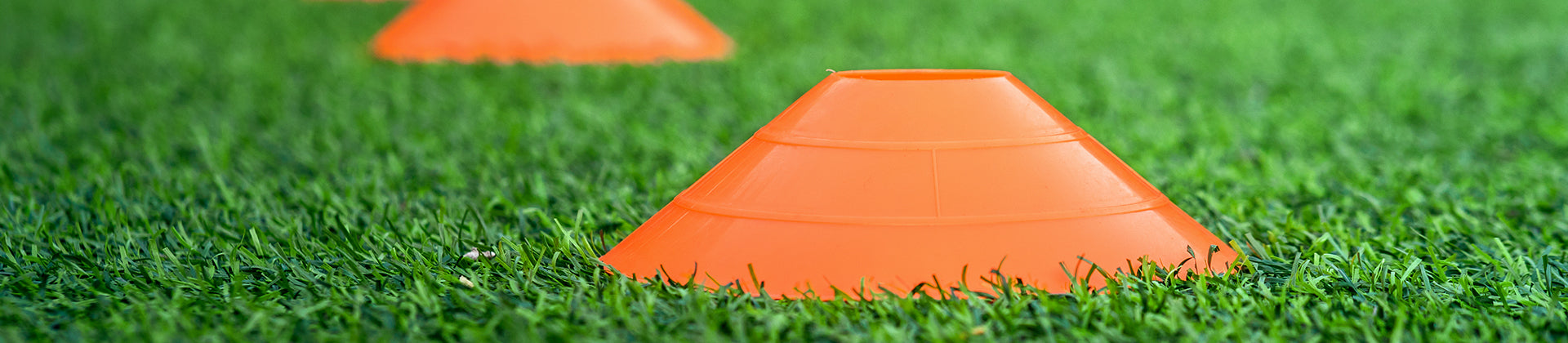 An orange plastic cone sits on green grass with another similar cone blurred in the background, commonly used for sports training or marking field boundaries.