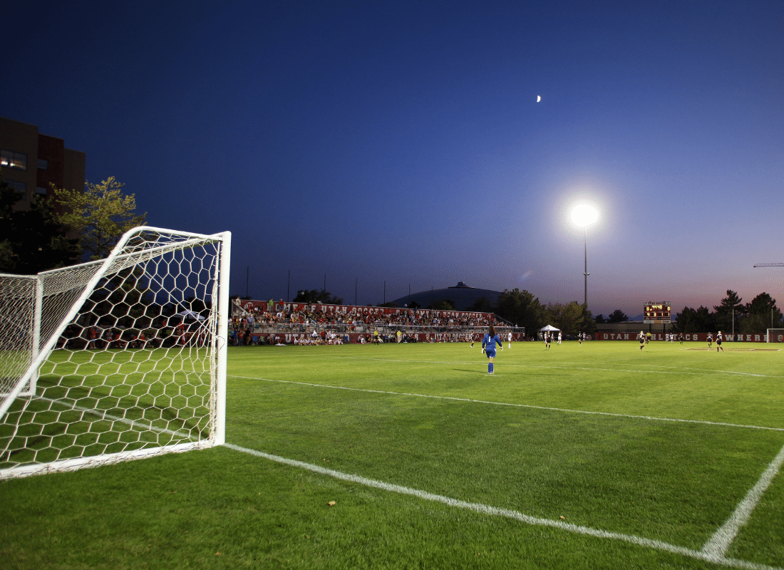 A soccer field at dusk with bright stadium lights on, a goalpost in the foreground, players on the field, and spectators seated in bleachers under a clear evening sky with a visible moon.