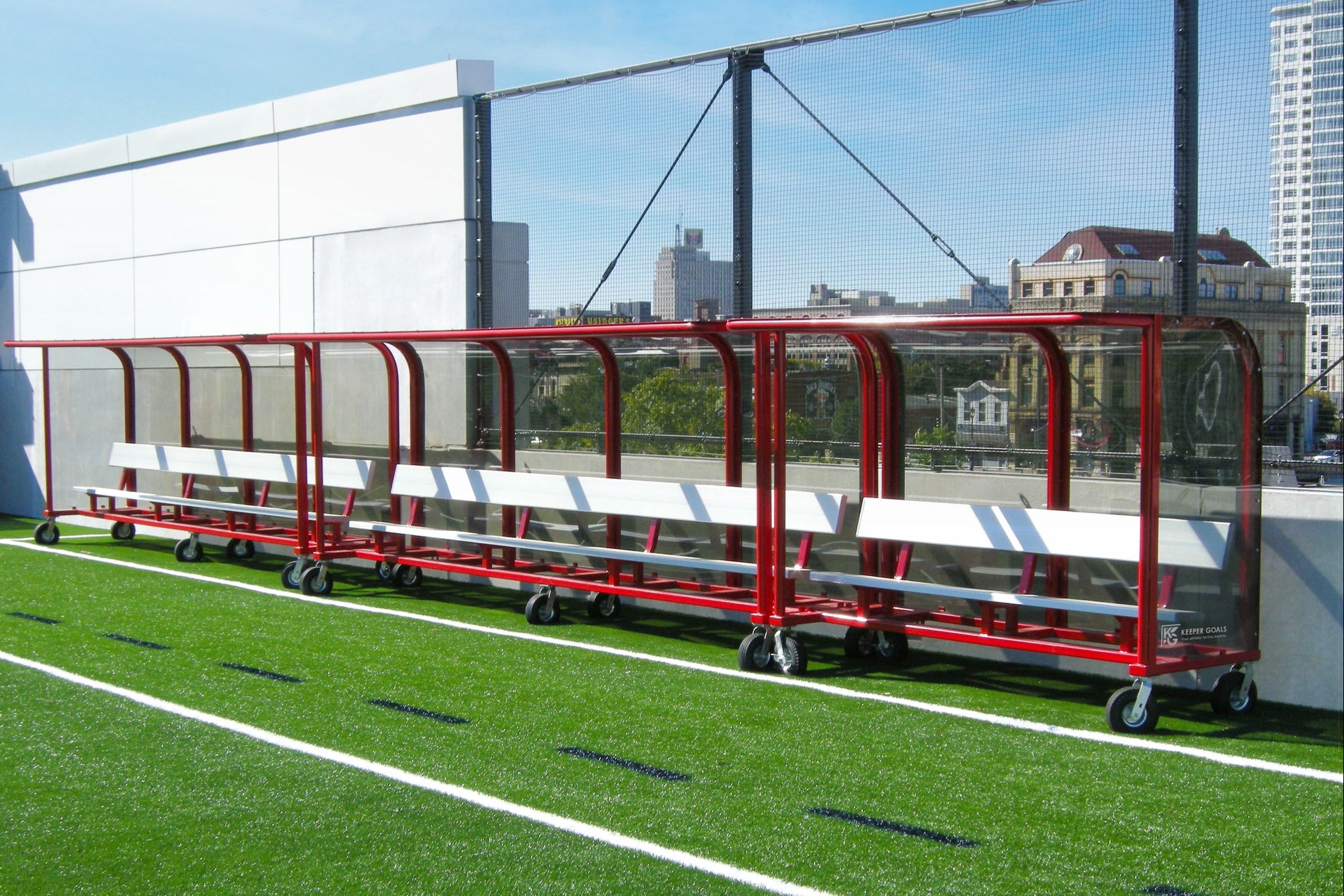 Two red and white covered team benches with wheels are positioned side by side on an outdoor artificial turf sports field, with a cityscape visible in the background behind a wire fence.