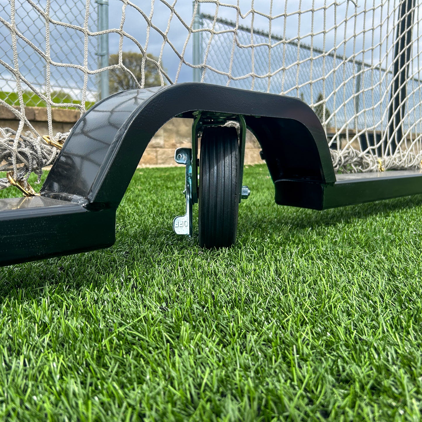A close-up of the wheel attachment on the Ultimate Wheeled Box-Style Soccer Goal positioned on artificial turf, showing the wheel beneath the frame with the net and a portion of the field visible in the background.