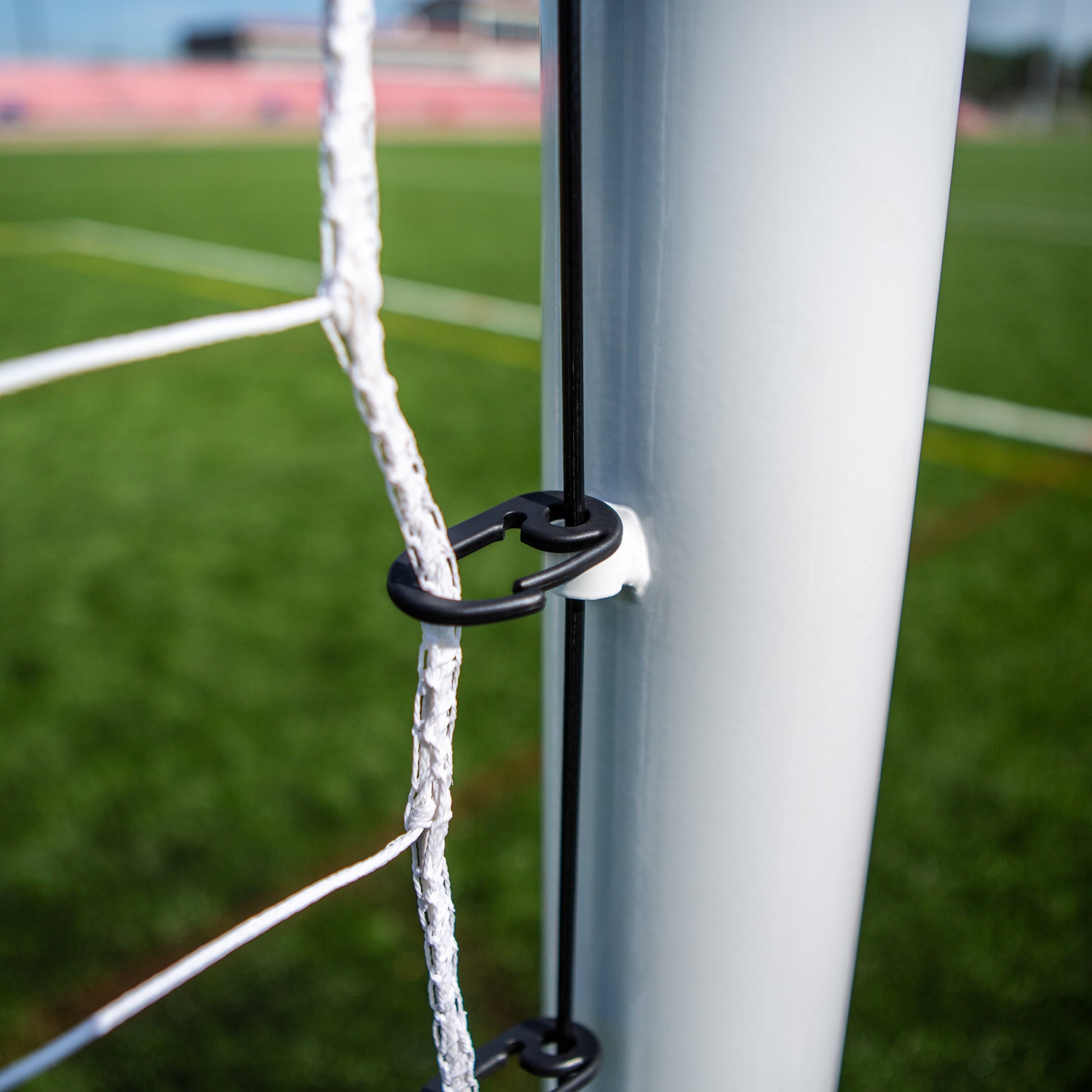 Close-up of the Ultimate Wheeled Box-Style Soccer Goal, showing black clips securing the net to the white metal post, with green artificial turf and a professional stadium goal blurred in the background.