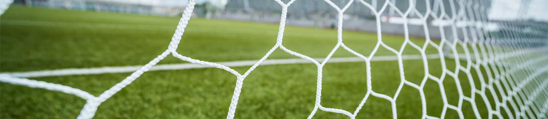 Close-up view of a white soccer goal net with a blurred green soccer field and white boundary lines in the background.