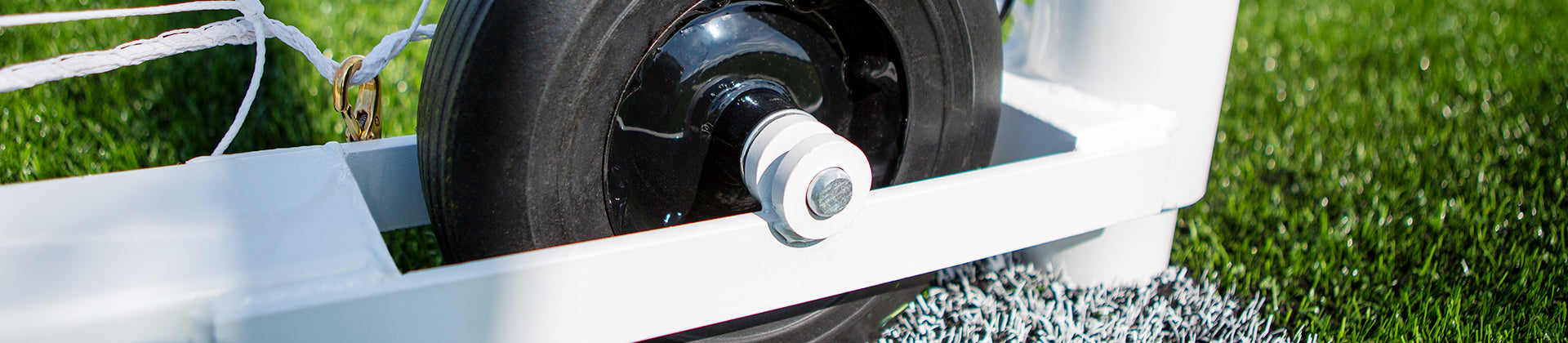 Close-up of a black wheel attached to the base of a white soccer goal, positioned on green artificial turf with white field markings visible.