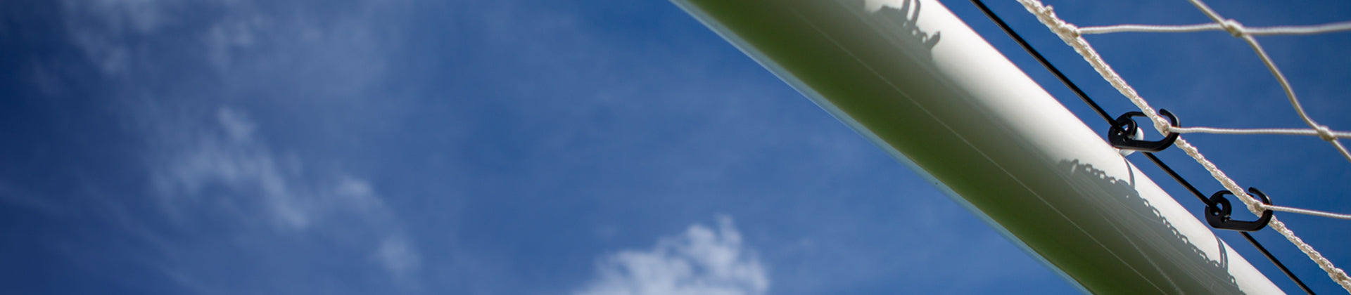 Close-up view of a volleyball net and pole against a bright blue sky with a few wispy clouds. The image is angled upward, emphasizing the net and sky.