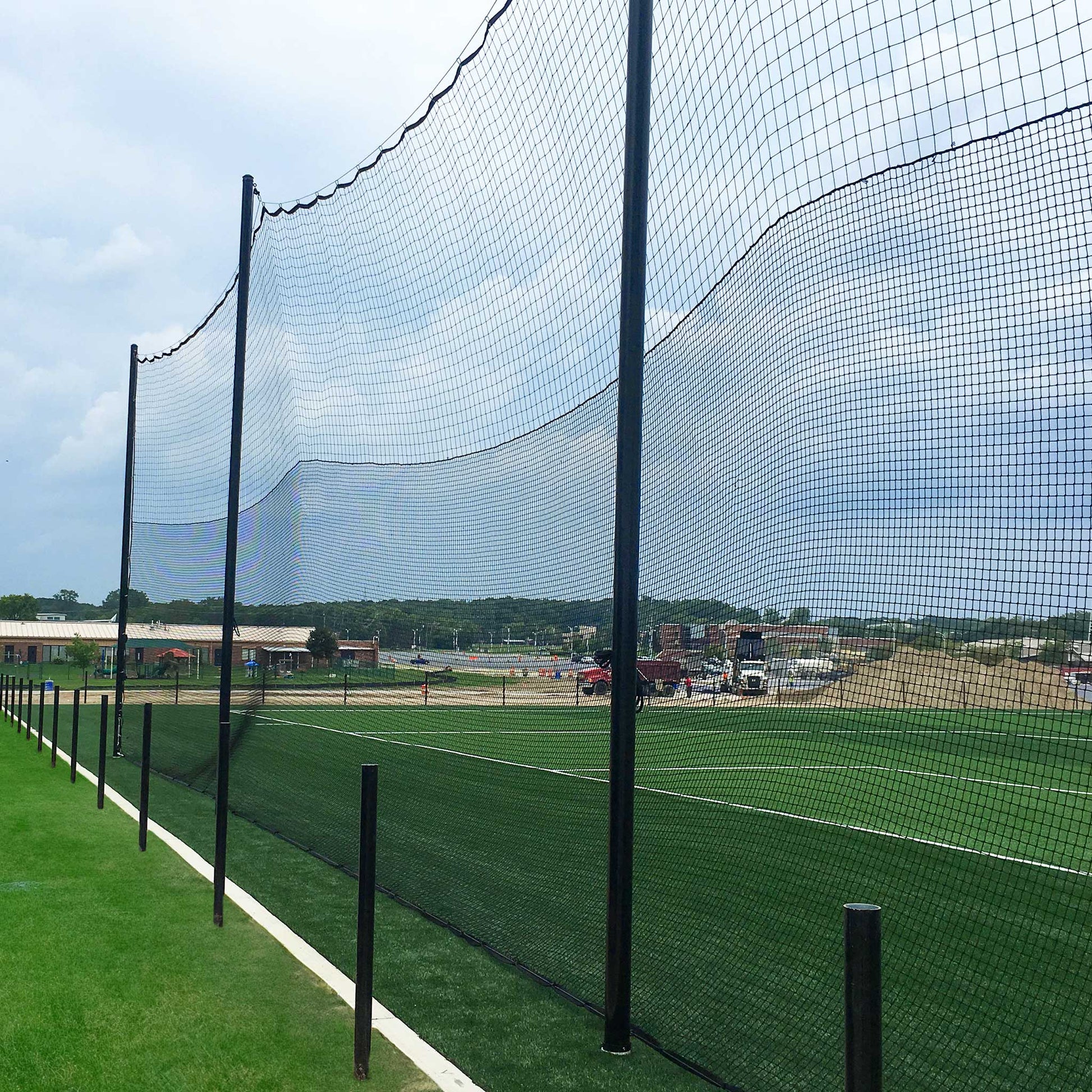 Hybrid Soccer & Lacrosse Backup Netting Systems with tall black netting and sturdy poles border a green artificial turf field, with buildings, vehicles, and a sandy construction area visible in the cloudy background.