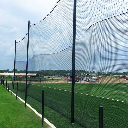 Hybrid Soccer & Lacrosse Backup Netting Systems with tall black netting and sturdy poles border a green artificial turf field, with buildings, vehicles, and a sandy construction area visible in the cloudy background.