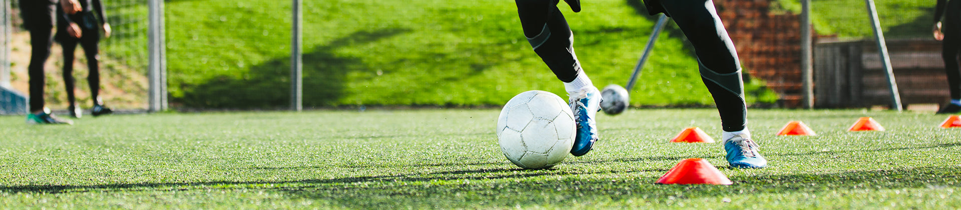 A soccer player dribbles a white ball around orange training cones on a green field during a practice session. Only the players legs, wearing black pants and blue shoes, are visible.