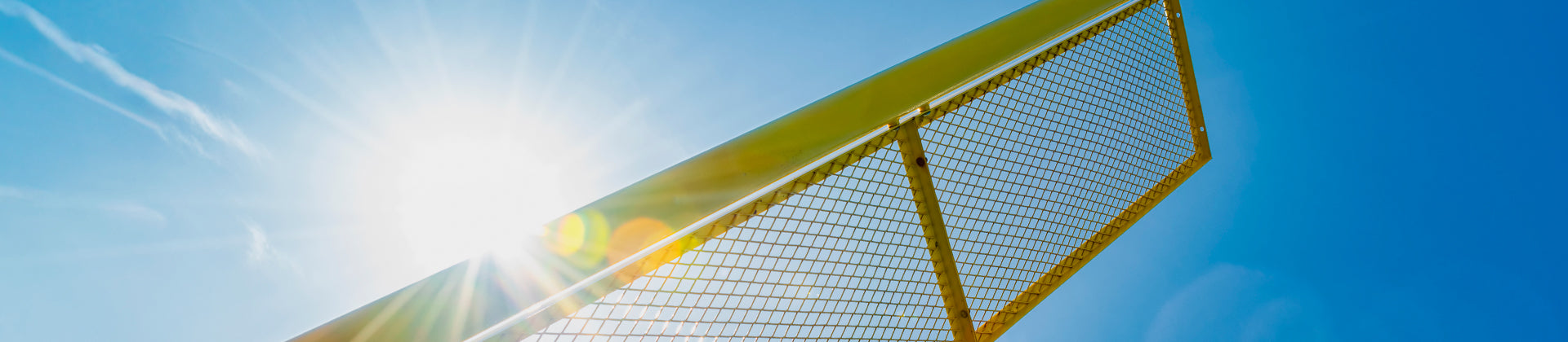 A yellow football goalpost photographed from below against a bright blue sky, with sunlight shining near the edge, creating a lens flare effect.