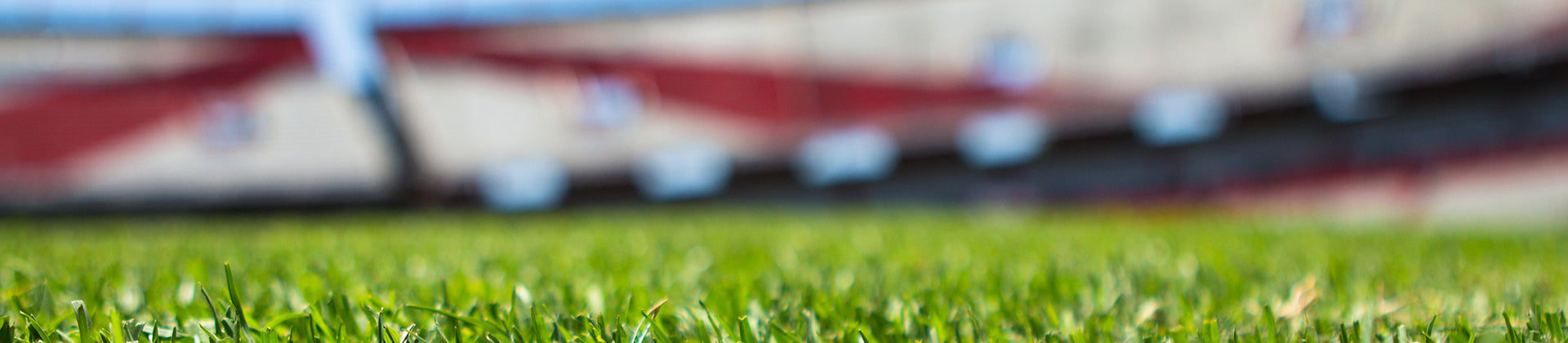 Close-up view of green grass on a sports field, with blurred stadium seats and structures in the background. The focus is on the grass in the foreground.