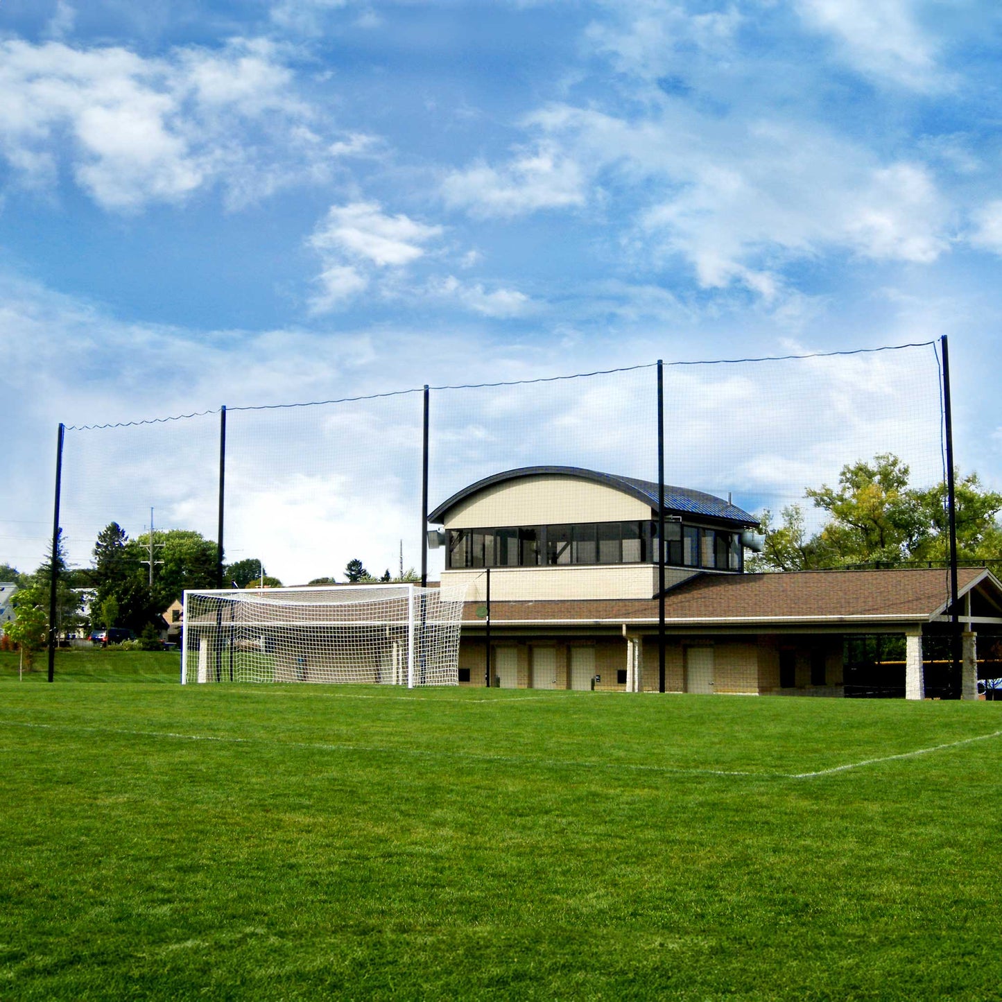 A soccer goal with aluminum posts is set on a grassy field, backed by Standard Soccer Backup Netting Systems. Behind, a building with a curved roof and trees are visible under a partly cloudy sky.