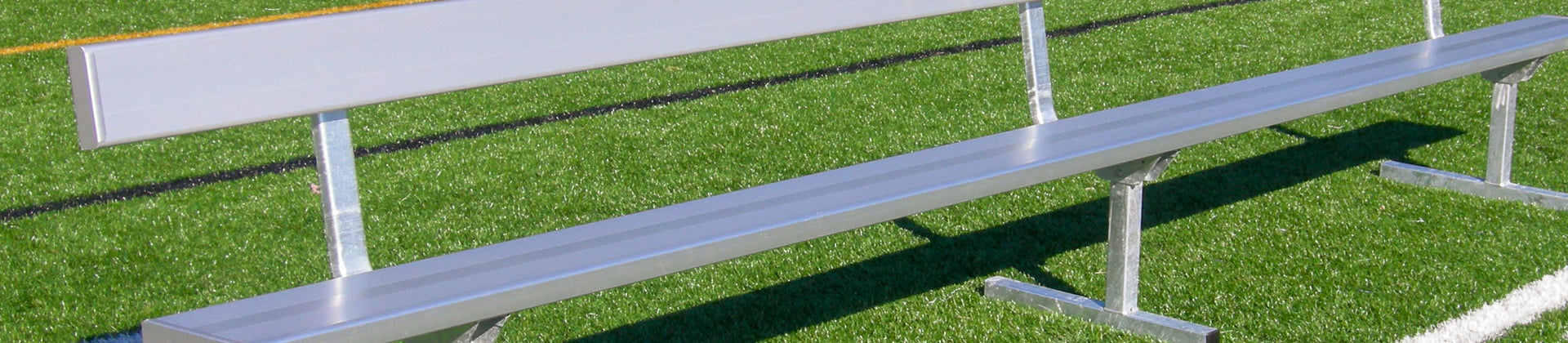 A long, silver metal bench with a backrest is positioned on green artificial turf, next to black and yellow field lines.
