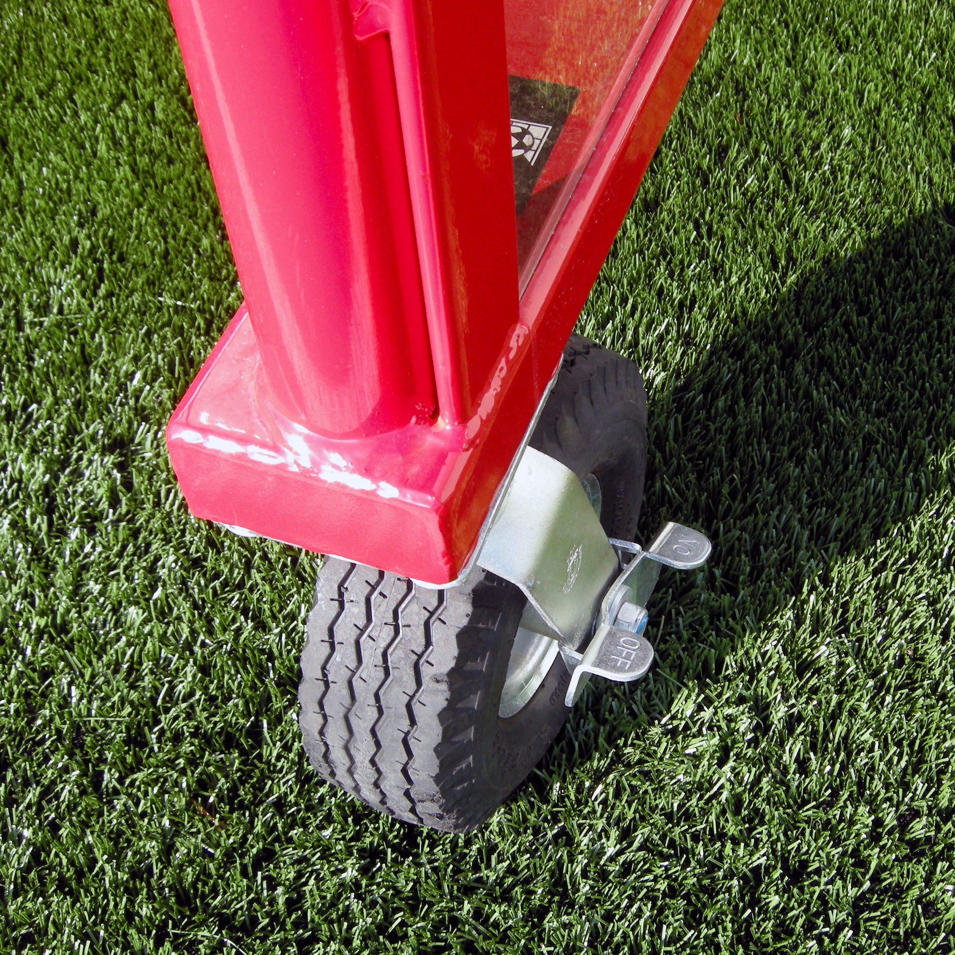 Close-up of a red cart’s corner with a large black rubber wheel on green artificial grass—perfect for moving equipment to the 20 Box-Style Heavy-Duty Team Shelter. The wheel includes OFF/ON metal foot pedals, likely for locking or unlocking.