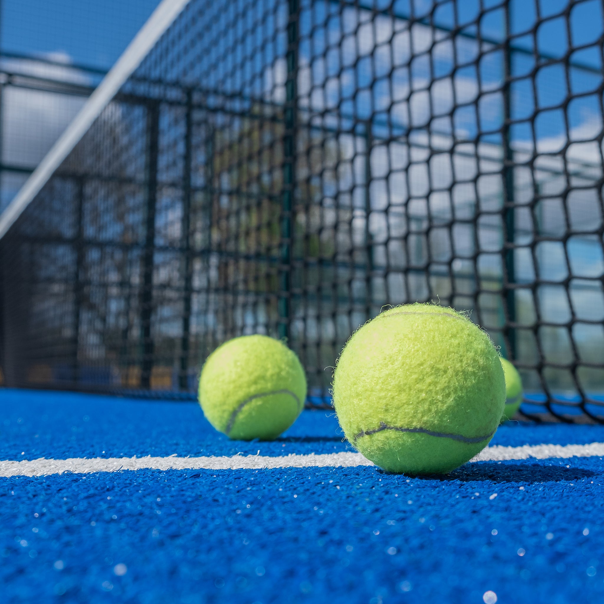 Close-up of three yellow tennis balls on a bright blue tennis court, with a net and blurred background under a sunny sky.