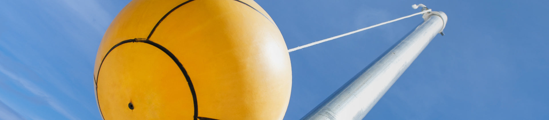 A close-up view of a yellow tetherball attached to a white rope, with the pole extending upwards against a clear blue sky.