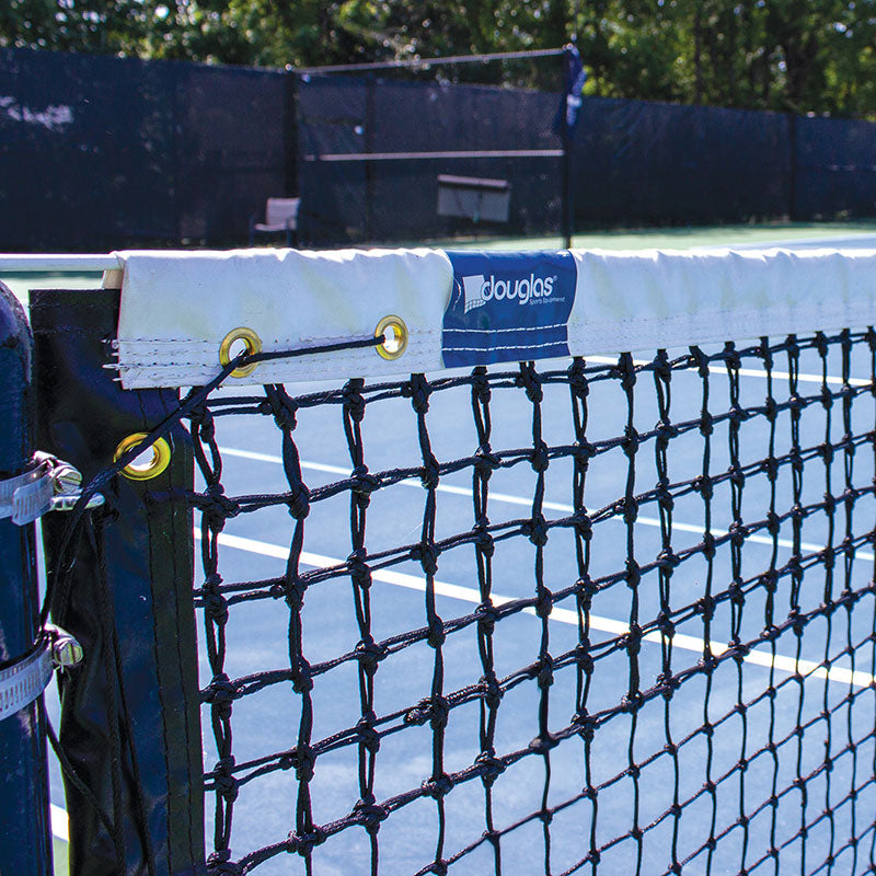 Close-up of the TN-28DM Double Mesh Tennis Net with braided polyethylene netting attached to a post on an outdoor court. White lines and trees are in the background, and the Douglas label is visible on the net’s white top band.