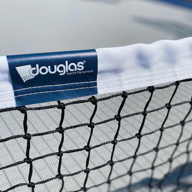 Close-up of the TN-45 Standard Tennis Net (Made in USA), a heavy-duty black net with a white top band and blue Douglas Sports Equipment tag. Meets USTA requirements. Blurred background hints at an outdoor tennis court.