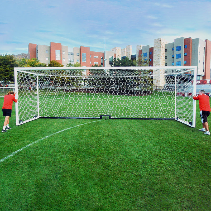 Two people in red shirts move an Ultimate Wheeled Box-Style Soccer Goal across a green field, with modern apartment buildings and trees visible in the background beneath a clear blue sky.