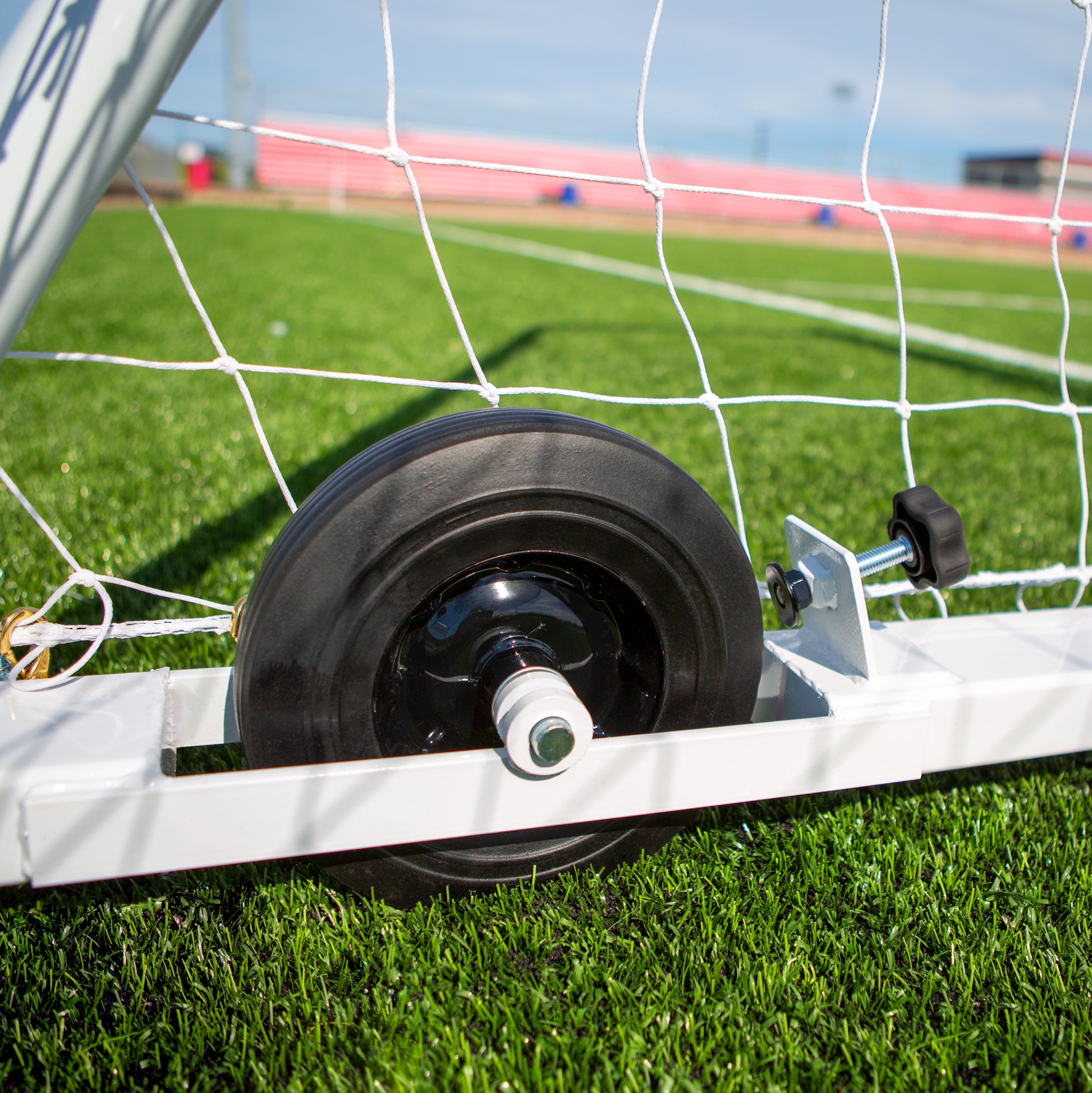 A close-up shows the black wheel of an Ultimate Wheeled Box-Style Soccer Goal attached to its white frame, set on green artificial turf with the goal net and a blurred outdoor sports field in the background.
