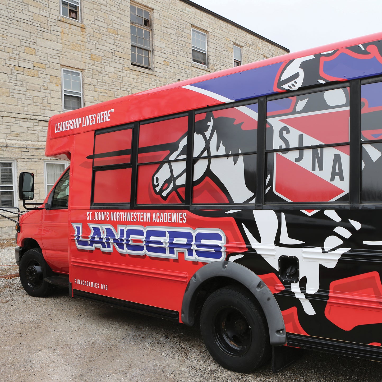 A red bus with Vehicle Wraps displays a white horse, SJNA crest, and “LANCERS,” plus the text St. Johns Northwestern Academies and Leadership Lives Here. The bus is parked beside a stone building.