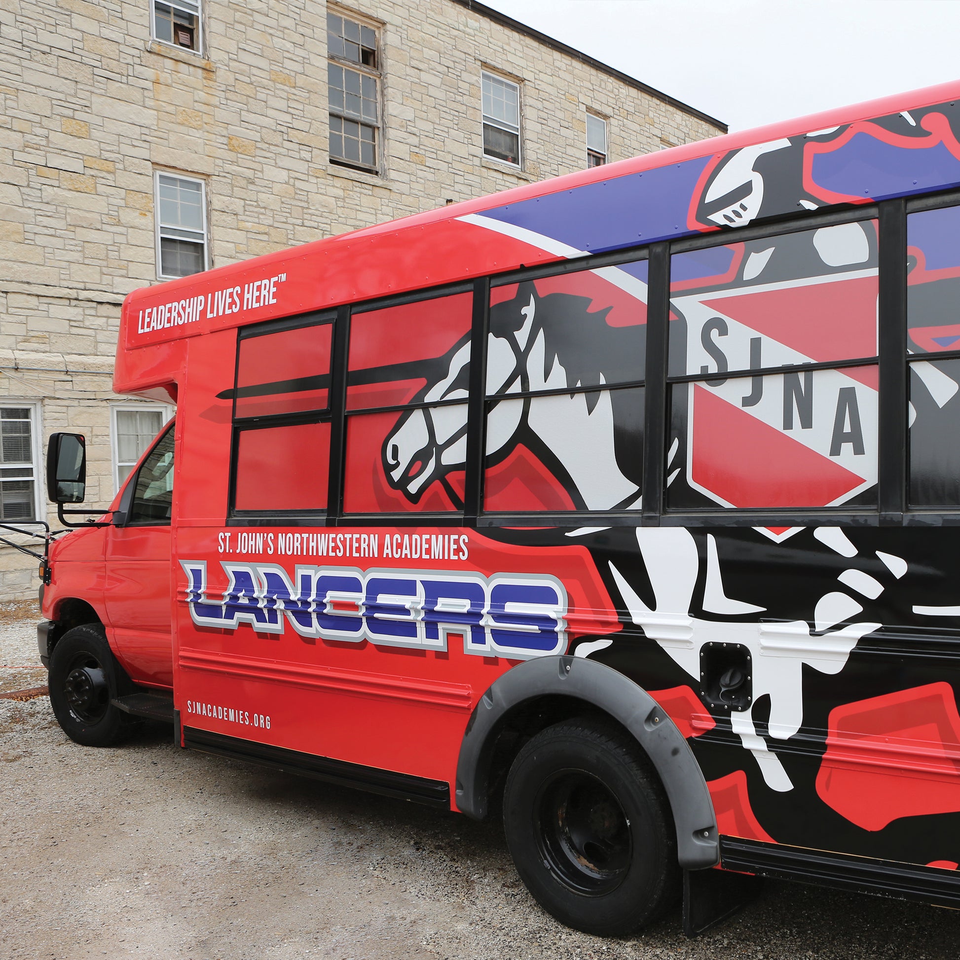 A red bus with Vehicle Wraps displays a white horse, SJNA crest, and “LANCERS,” plus the text St. Johns Northwestern Academies and Leadership Lives Here. The bus is parked beside a stone building.
