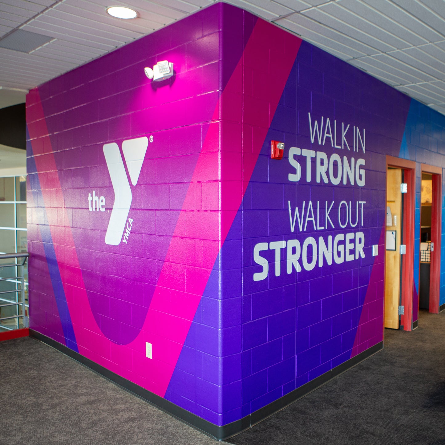 A Vinyl Wall Wrap at a YMCA displays the Y logo and WALK IN STRONG WALK OUT STRONGER in bold white letters, covering interior walls with custom shades of purple, pink, and blue.