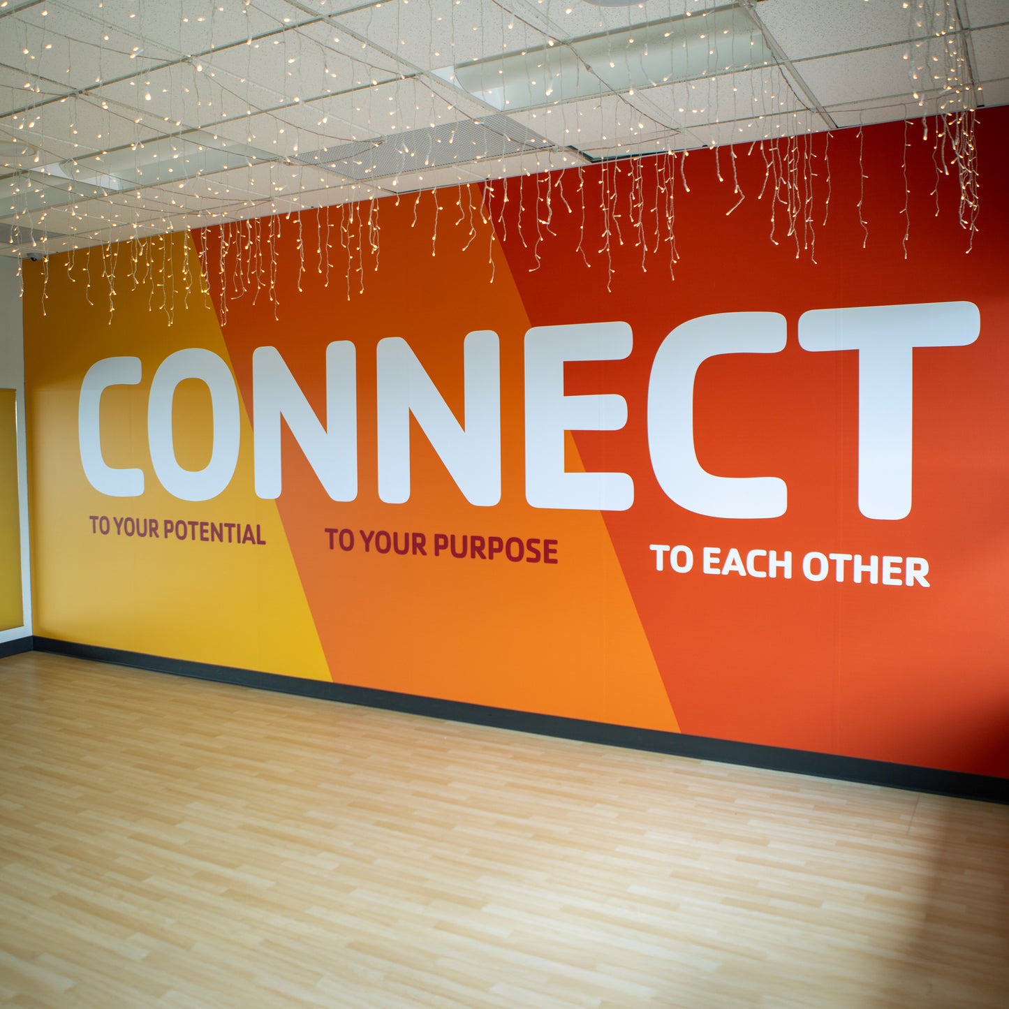 A Vinyl Wall Wrap with a vibrant orange-yellow gradient displays bold white text: CONNECT, with smaller lines reading TO YOUR POTENTIAL, TO YOUR PURPOSE, and TO EACH OTHER. String lights hang overhead for added ambiance.