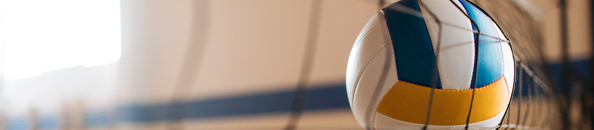 A volleyball with blue, yellow, and white panels hitting or passing through a volleyball net inside a gymnasium.