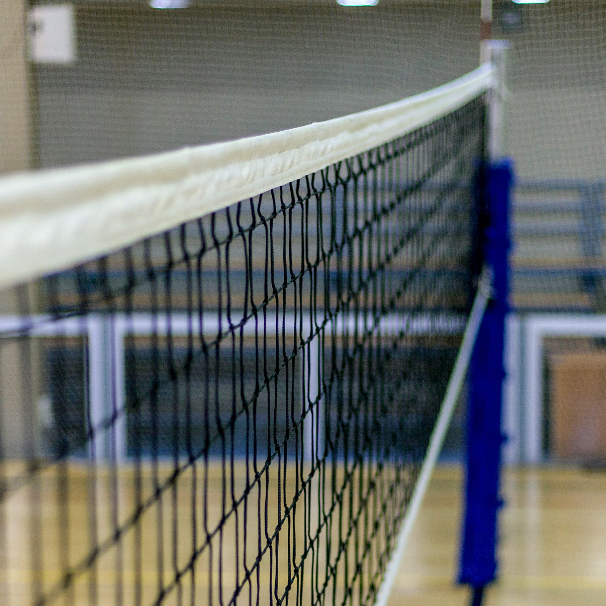 Close-up view of an indoor volleyball net stretching across a wooden gym floor, with blurred bleachers and sports equipment visible in the background.
