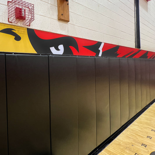 A Wall Pad in black poly-foam covers the lower gym wall. Above, a mural in red, yellow, and black shows part of a fierce animal face. A red fire alarm is also mounted on the wall.
