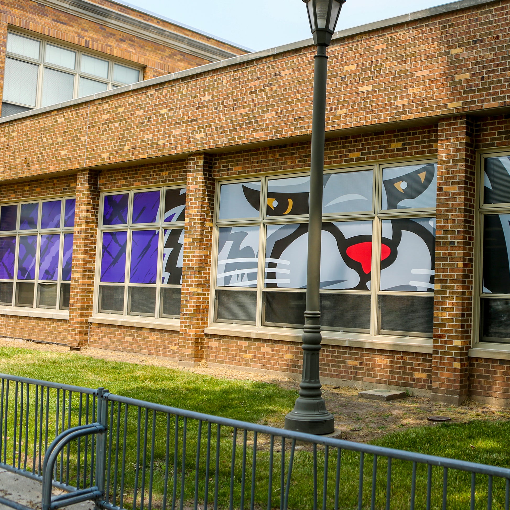 A vibrant large cat face mural with bold colors and sharp lines is printed on Perforated Window Film, spanning multiple windows of a brick building to provide privacy. A metal fence and lamppost stand before the colorful artwork.
