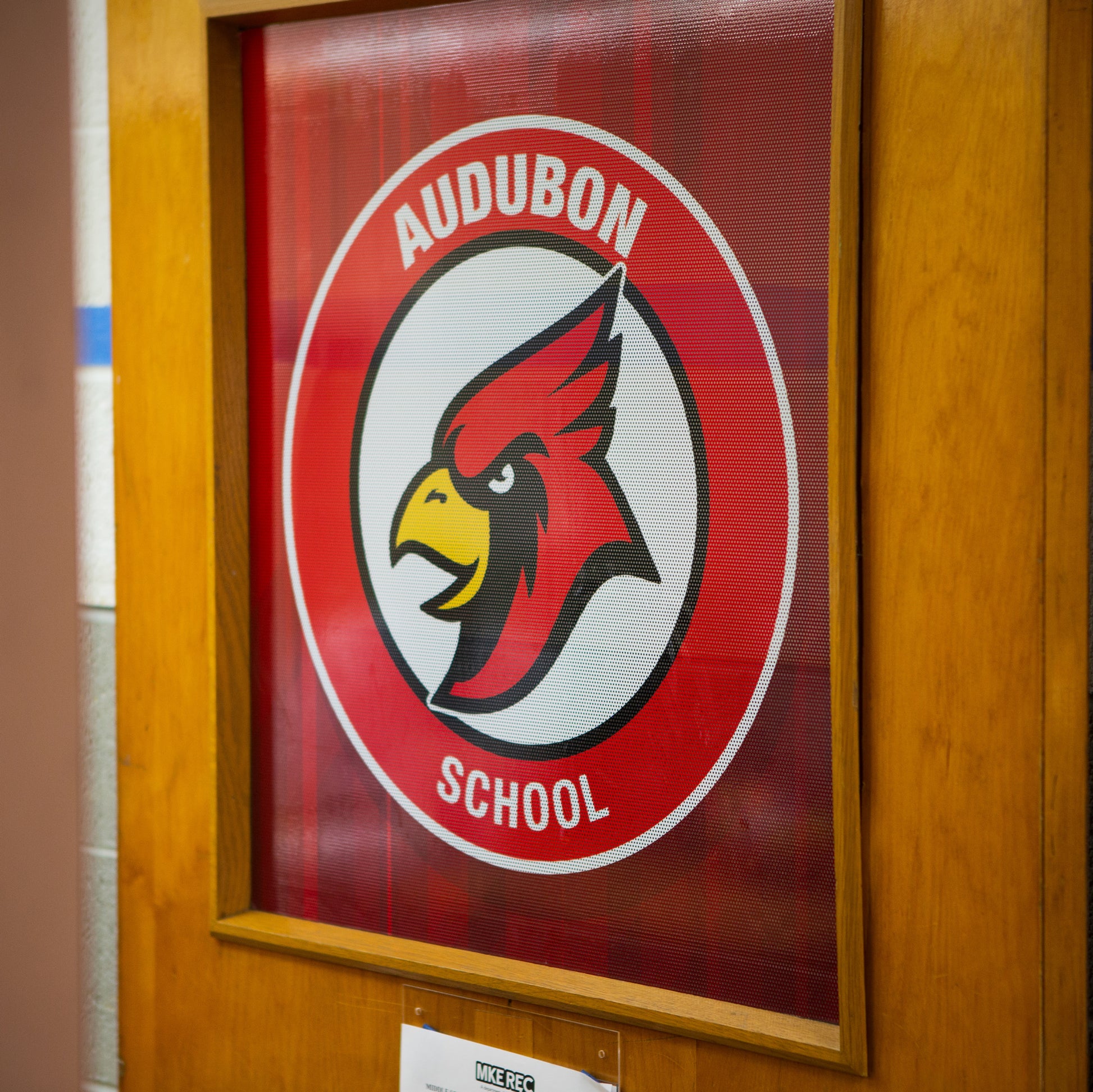 Perforated Window Film on a wood-framed glass door displays a circular red cardinal head logo, encircled by Audubon School in white letters on a red background. It offers privacy and highlights your brand.