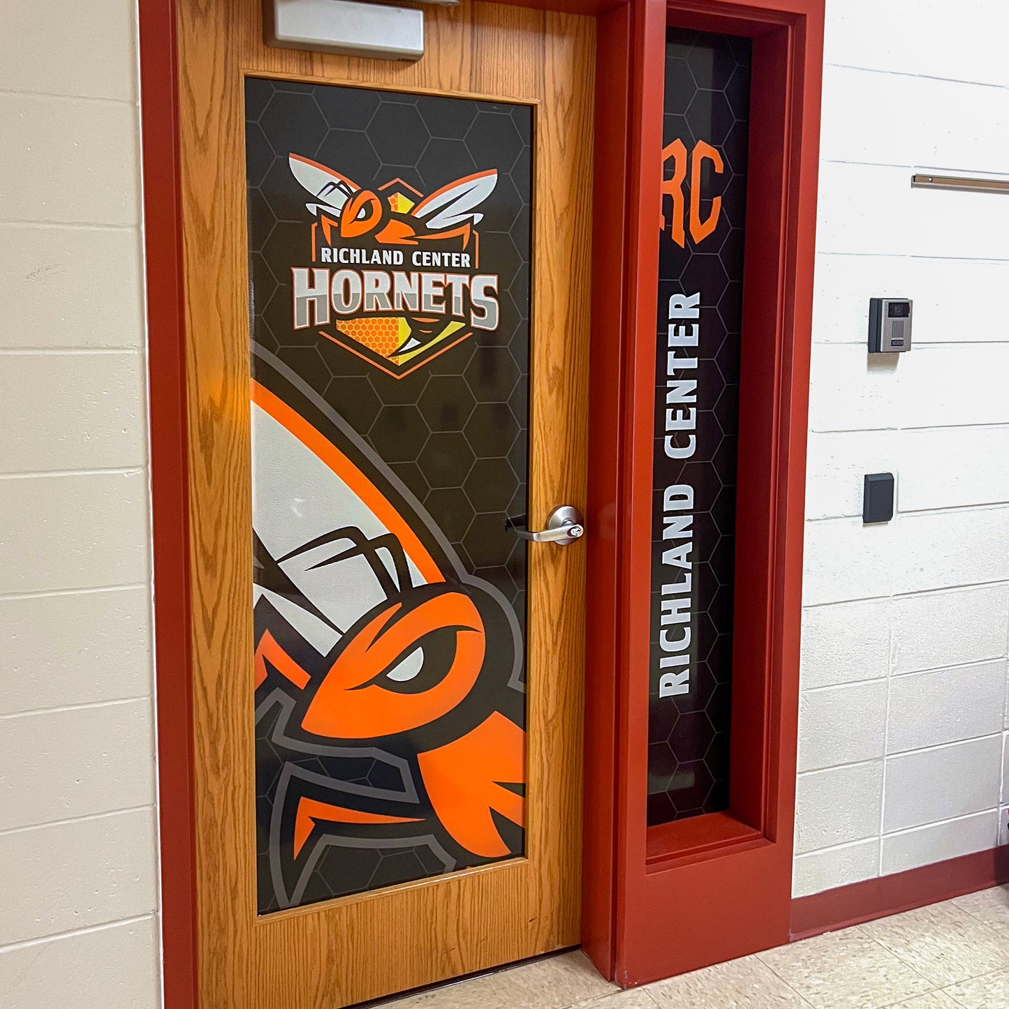 A wooden door displays a large hornet graphic and Richland Center Hornets on a honeycomb background. The side window uses Perforated Window Film, offering privacy protection and showcasing your unique branding.