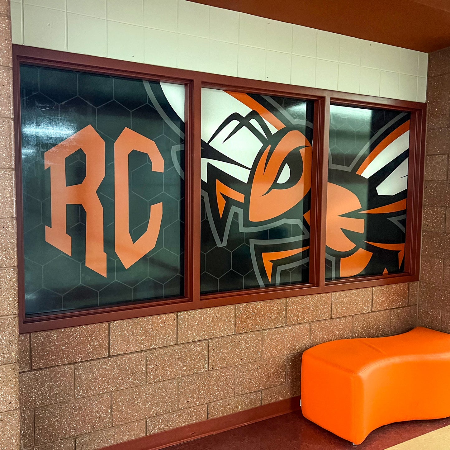 A large wall graphic shows orange RC letters and a hornet mascot, framed by three window panes fitted with Perforated Window Film for privacy, above an orange curved bench against brown and white tiled walls.