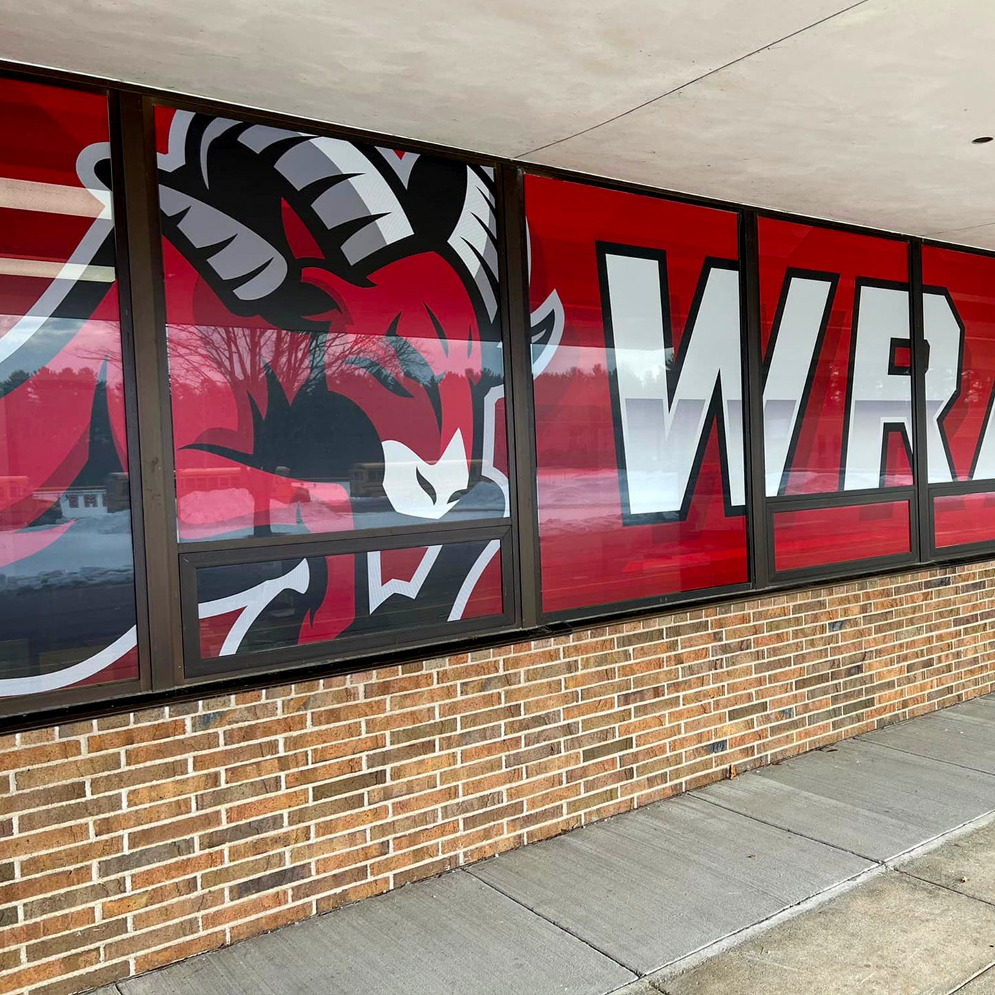 A building window displays an angry red ram mascot and white WRA letters on a red background using Perforated Window Film, providing striking branding and enhanced privacy.