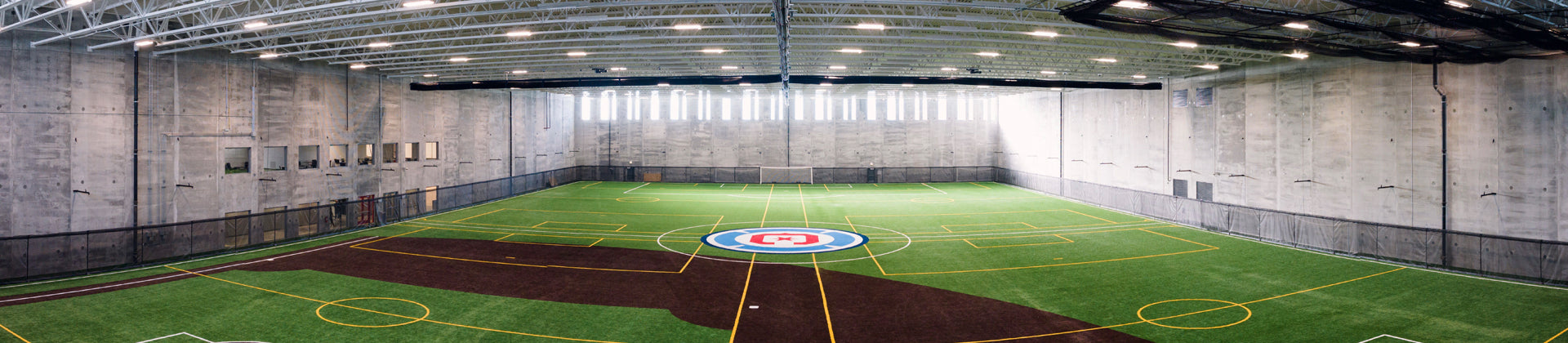 Wide-angle view of an indoor sports facility with artificial turf, marked for multiple sports like soccer and lacrosse, surrounded by high concrete walls and illuminated by rows of ceiling lights.