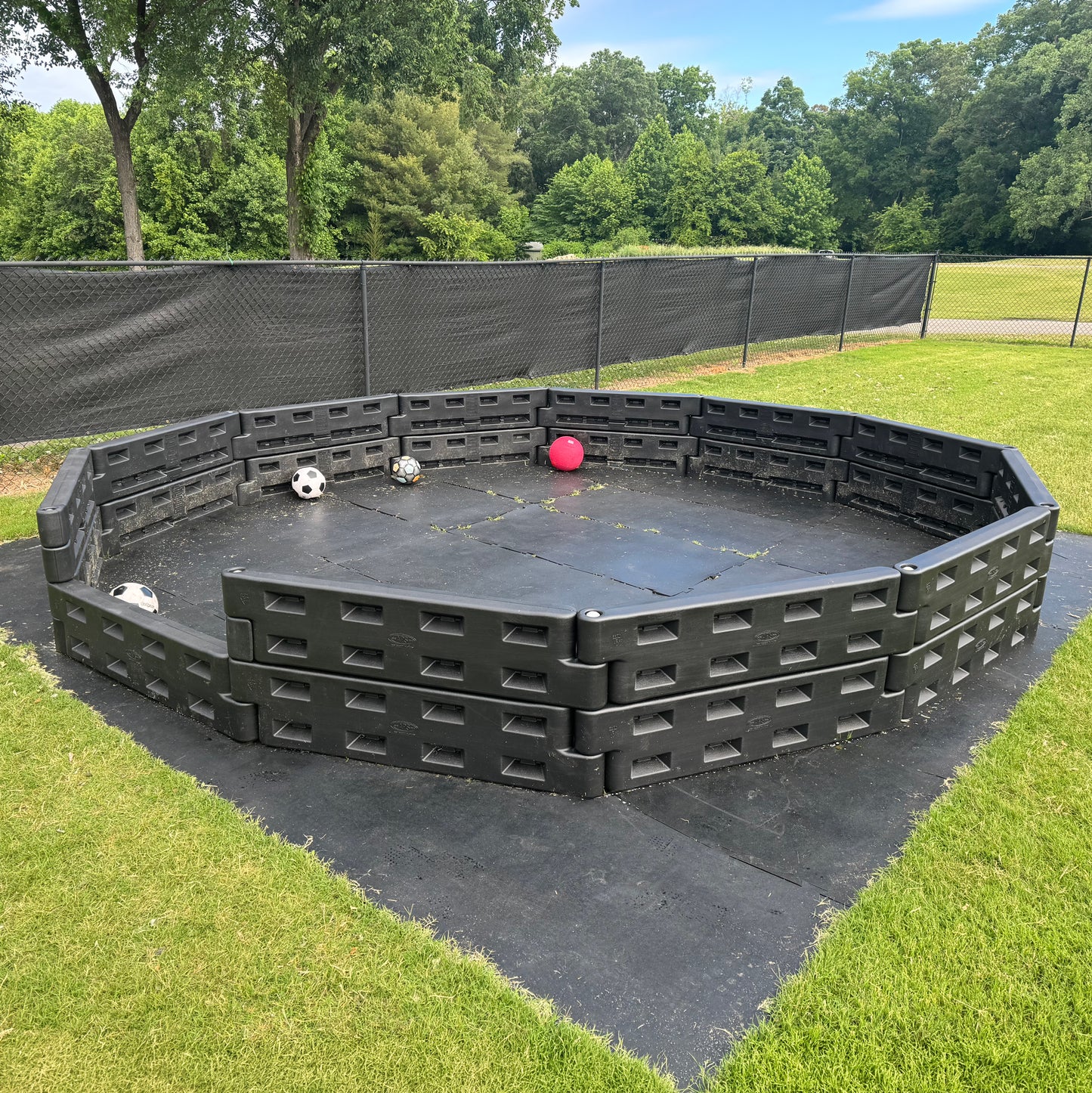 An outdoor playground features the APS Standard GaGa Ball Pit | Permanent In-Ground Mount with black plastic walls on grass, holding several balls including two soccer balls and a red ball. Trees and a chain-link fence are in the background.