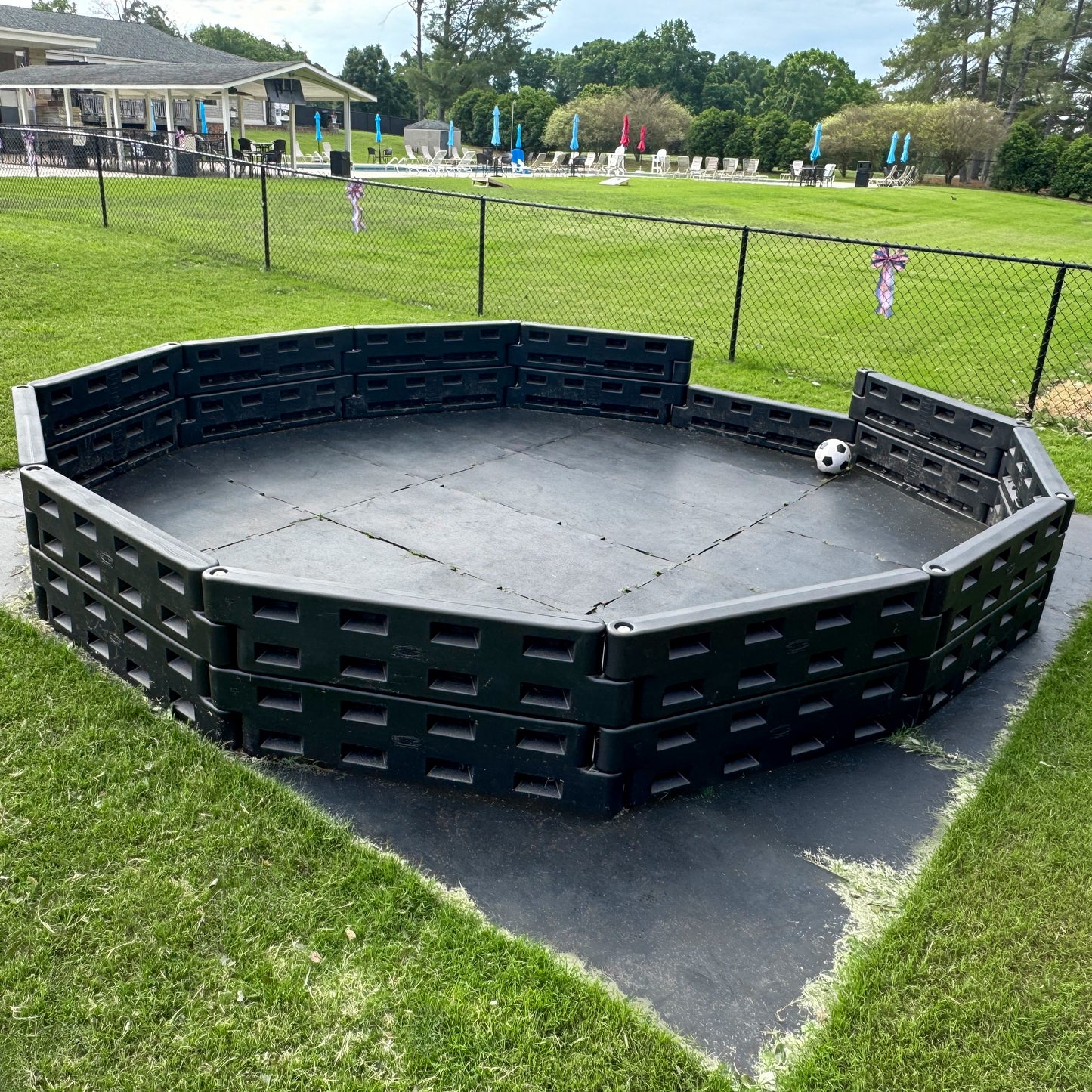 An APS Standard GaGa Ball Pit | Permanent In-Ground Mount with sturdy black plastic walls is installed on grass near a fenced pool and building, with a white and black ball resting inside against the wall.