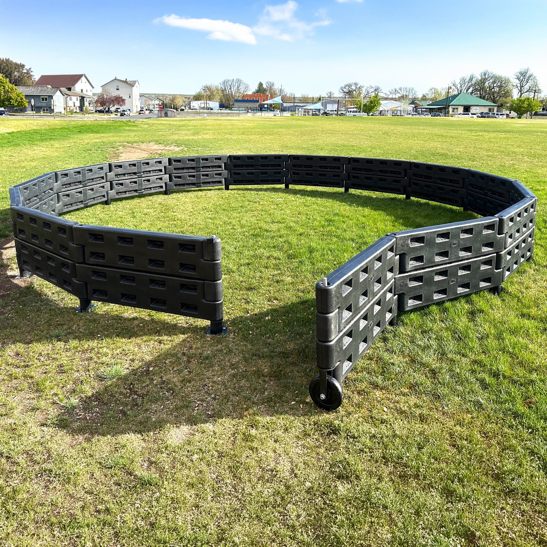 The APS Premium Gaga Ball Pit with ADA Gate, made of black plastic in an octagonal shape, is set up on grass under a blue sky with buildings and trees in the background, ready for playground games.