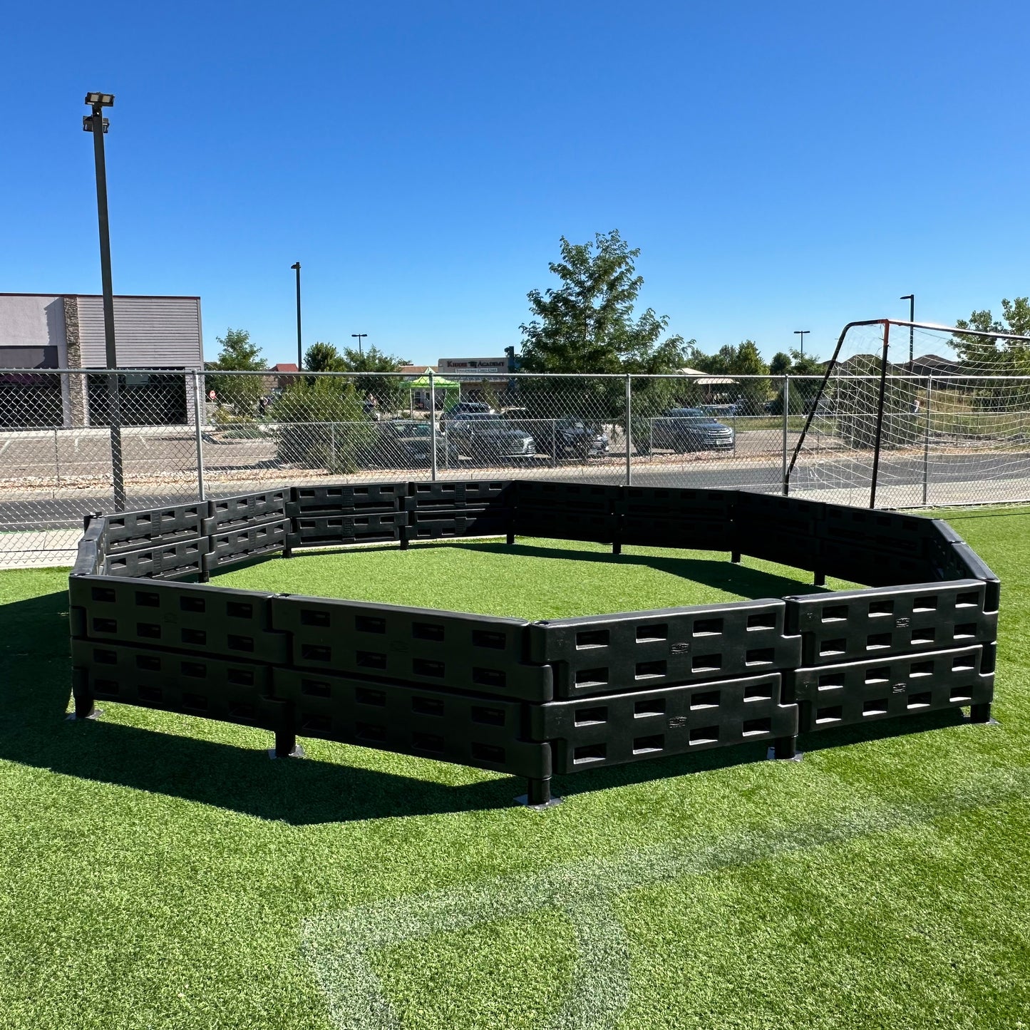 The APS Premium Gaga Ball Pit with ADA Gate, made of black plastic in an octagonal shape, sits on green artificial turf outdoors near a chain-link fence, with buildings, trees, and a clear blue sky in the background.