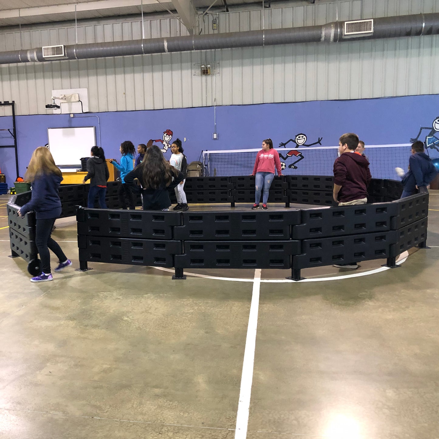 Children play gaga ball inside the APS Premium Gaga Ball Pit with ADA Gate in a gym with blue walls and cartoon murals, enjoying this popular playground game while others watch from outside the pit.