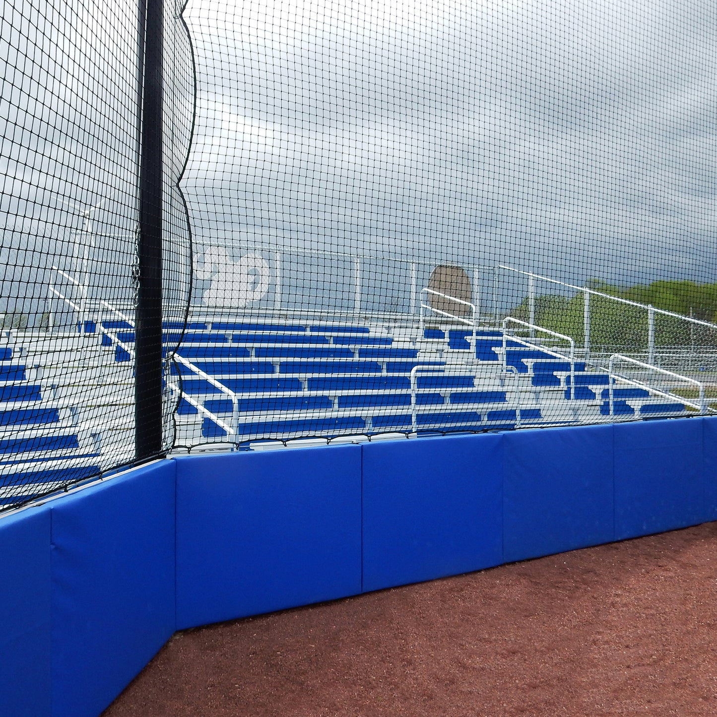 Empty blue bleachers behind a protective net at a baseball or softball field, under cloudy skies. The seating area has metal railings and is enclosed by Outdoor Protective Wall and Fence Padding made of durable vinyl panels.