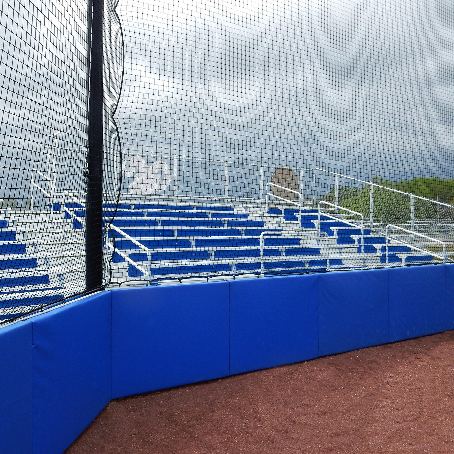 Empty blue bleachers behind a protective net at a baseball or softball field, under cloudy skies. The seating area has metal railings and is enclosed by Outdoor Protective Wall and Fence Padding made of durable vinyl panels.