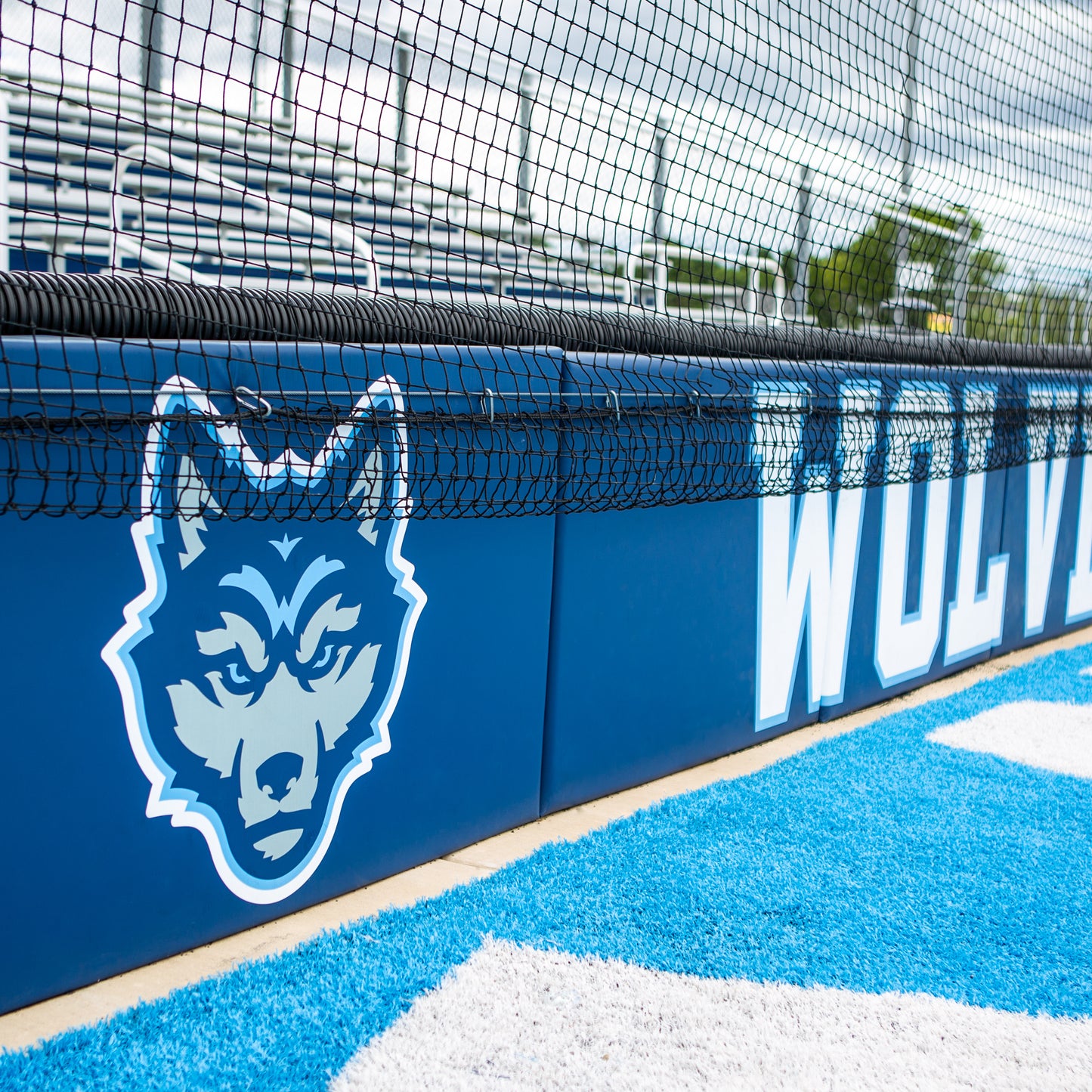 The Outdoor Protective Wall and Fence Padding features a blue and gray wolf mascot logo with the word WOLVES partially visible. Vinyl panels, a net, and bleachers appear in the sports field background.