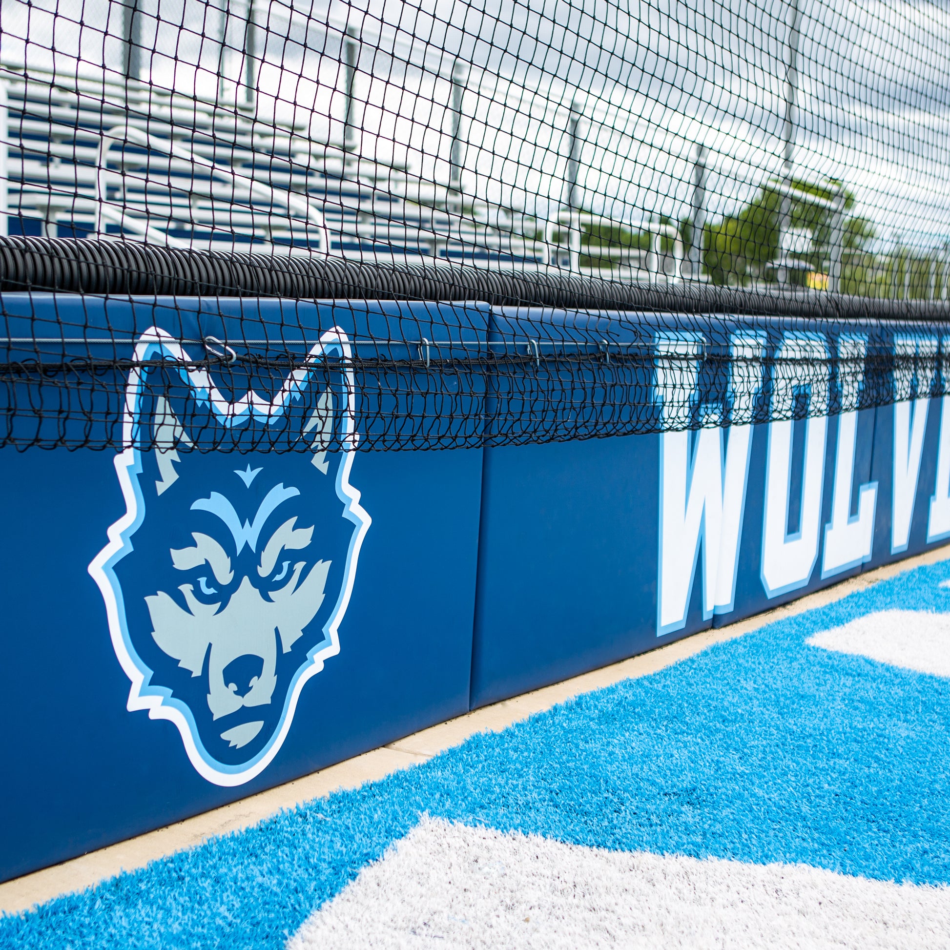 The Outdoor Protective Wall and Fence Padding features a blue and gray wolf mascot logo with the word WOLVES partially visible. Vinyl panels, a net, and bleachers appear in the sports field background.