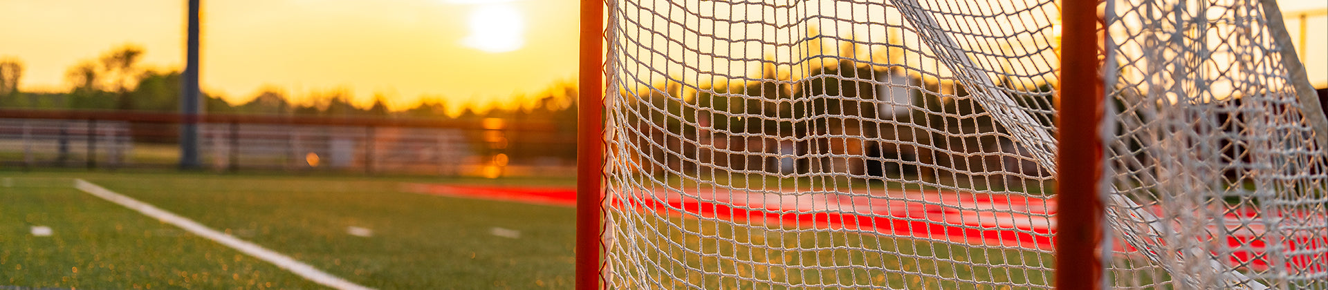 A close-up of a lacrosse goal net on a field at sunset, with the sun low in the sky, casting a warm golden glow over the green turf and distant trees.