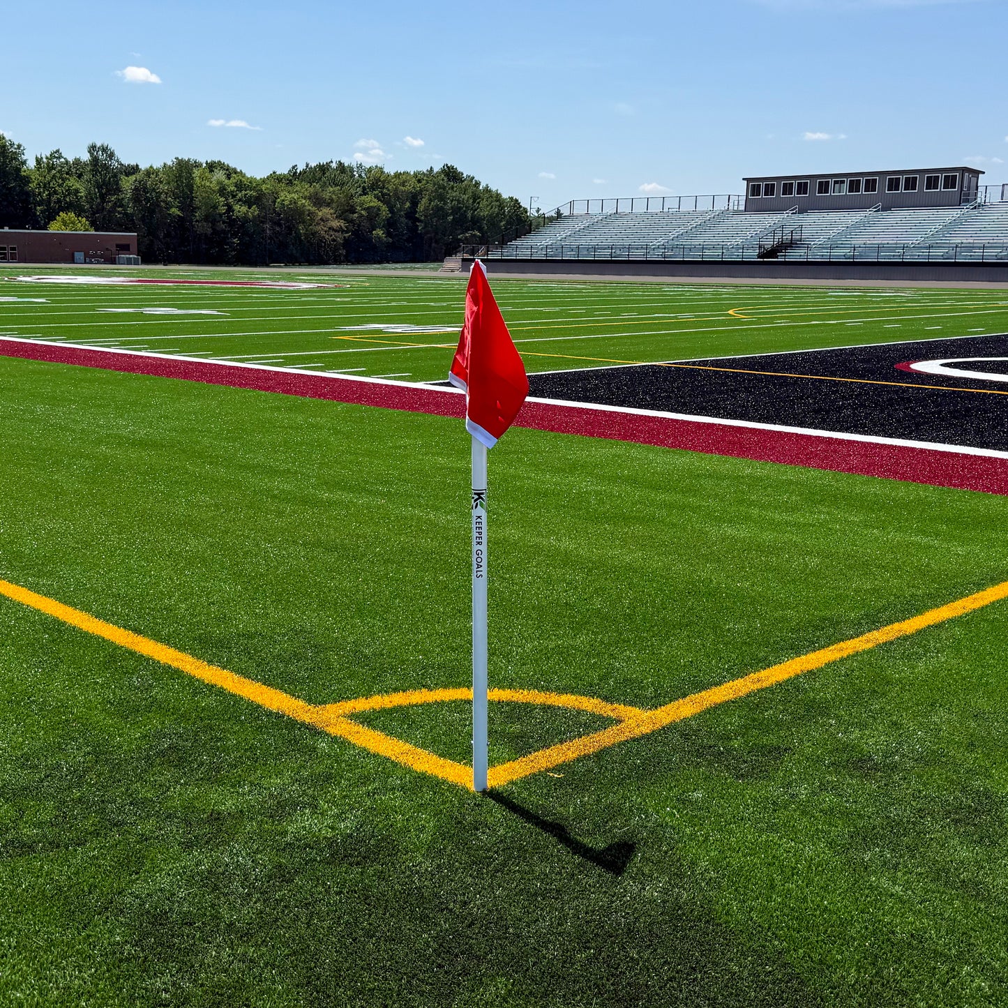 A set of Keeper Goals Sleeved Official Soccer Corner Flags, featuring a vibrant red flag, stands at the intersection of yellow soccer field marking lines on a green sports field, with bleachers and trees visible in the background under a clear sky.