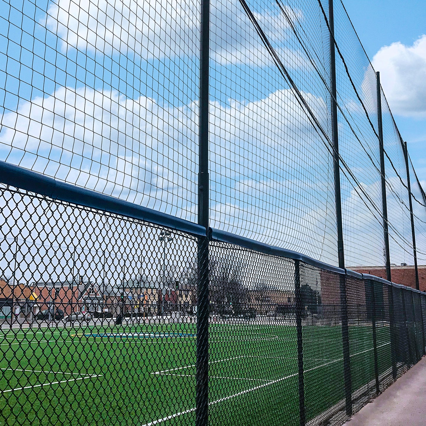 A green sports field at a multi-sport facility is enclosed by a tall chain-link fence under partly cloudy skies. Custom Engineered Backstop and Backup Netting Systems offer added protection, with buildings visible in the background.