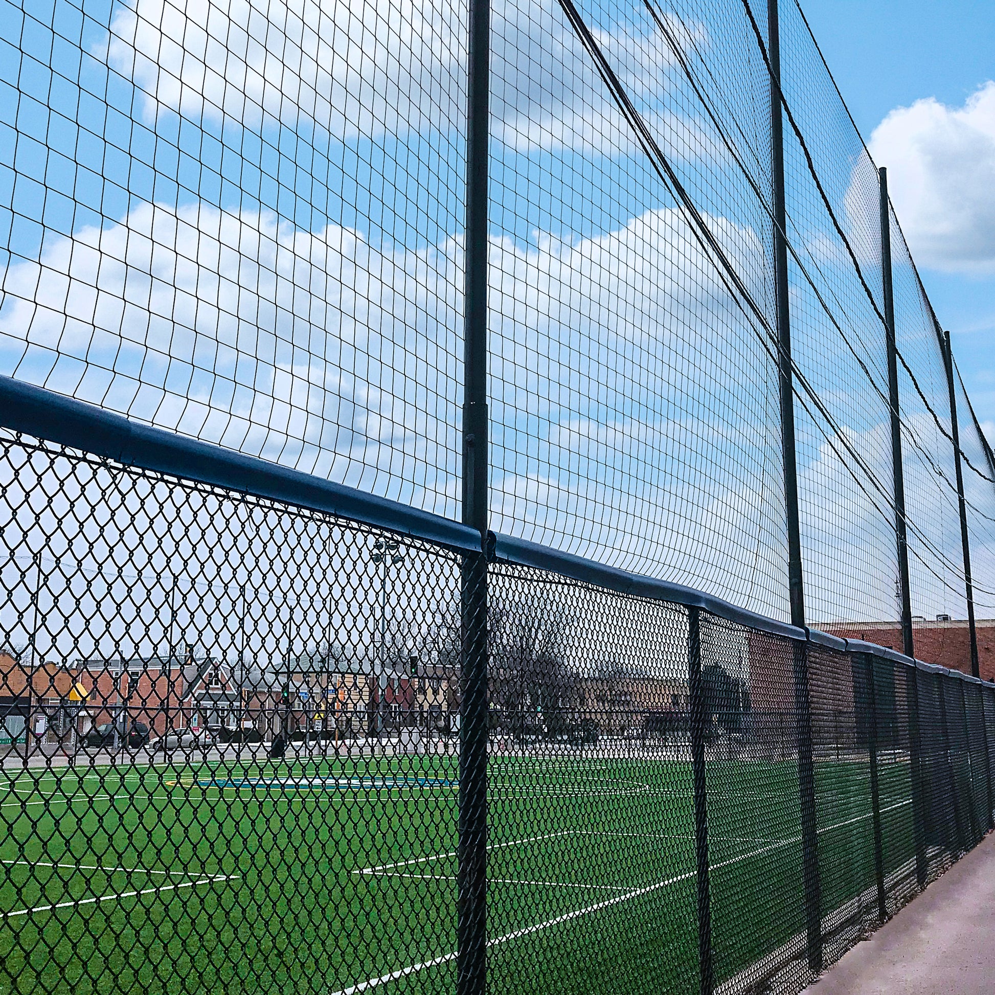 A green sports field at a multi-sport facility is enclosed by a tall chain-link fence under partly cloudy skies. Custom Engineered Backstop and Backup Netting Systems offer added protection, with buildings visible in the background.