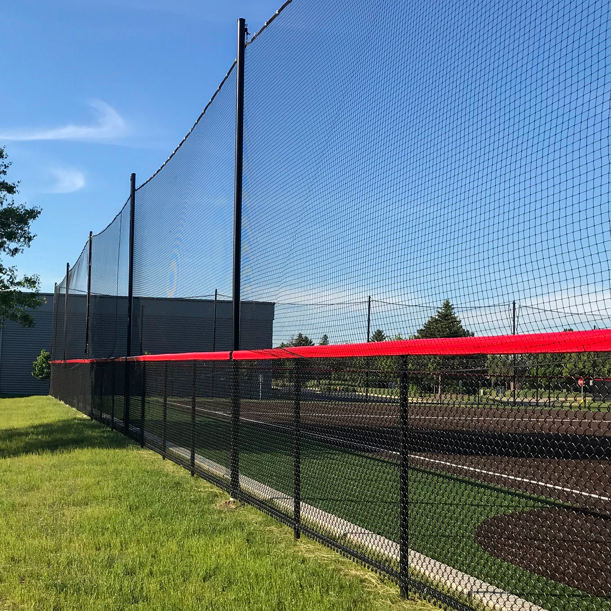 A Custom Engineered Backstop and Backup Netting System with a red stripe lines the grassy area and athletic field, featuring durable chain-link fencing beneath a clear blue sky.