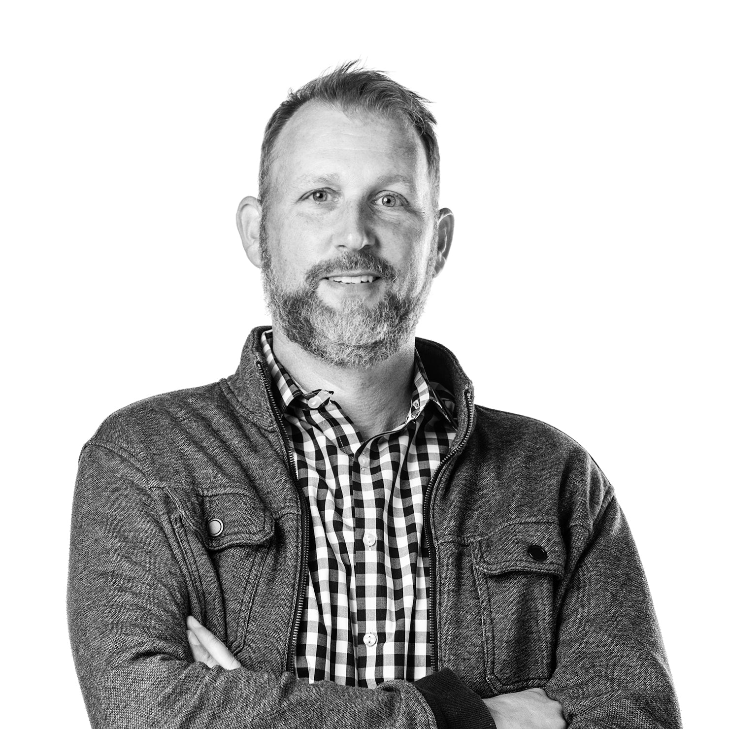 A man with short hair and a beard stands with arms crossed, wearing a checkered shirt and a zip-up jacket, looking confidently at the camera against a plain white background.